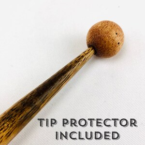May include: A wooden hair stick with a round, ball-shaped top and a tapered shaft. The stick is a warm brown color with darker wood grain patterns. The text "TIP PROTECTOR INCLUDED" is printed in black below the stick.