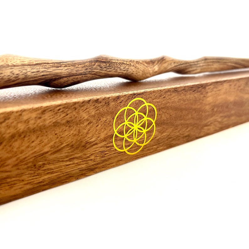 May include: A wooden wand with a natural, textured finish rests on a rectangular wooden box. The box features a yellow, stylized flower design. The wand has a curved, organic shape.