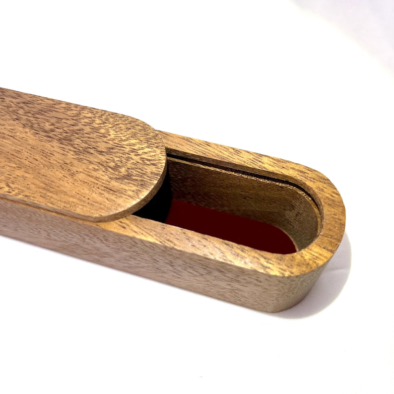 May include: A wooden box with a sliding lid, partially open to reveal a red interior. The box is made of light brown wood with a visible grain pattern. The box is oval-shaped.