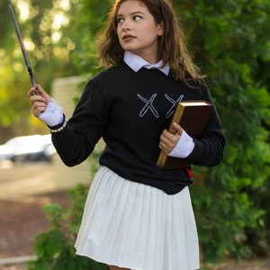 Puede incluir: Una persona con una sudadera negra con cruces bordadas blancas, una camisa blanca con cuello y una falda plisada blanca. Sostiene una varita y un libro marr&oacute;n con ribetes dorados. El atuendo tiene una est&eacute;tica de uniforme escolar.