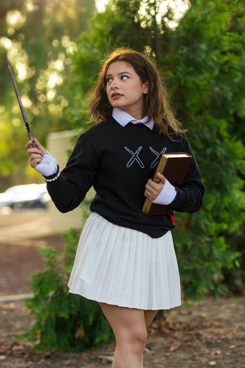 Puede incluir: Una persona con una sudadera negra con cruces bordadas blancas, una camisa blanca con cuello y una falda plisada blanca. Sostiene una varita y un libro marr&oacute;n con ribetes dorados. El atuendo tiene una est&eacute;tica de uniforme escolar.
