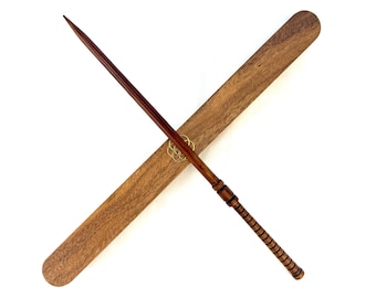 Puede incluir: Una varita de madera marrón oscuro con punta y mango detallado, colocada en diagonal sobre una funda de madera a juego. La funda tiene bordes redondeados y un emblema dorado. La varita mide aproximadamente 35,5 cm de largo.