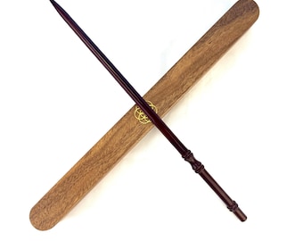 Puede incluir: Una varita marrón oscuro con punta afilada y mango adornado descansa en diagonal sobre una caja de madera. La caja es rectangular con bordes redondeados y presenta un emblema dorado. La varita mide aproximadamente 35 cm de largo.