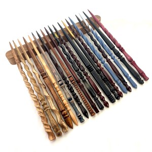 May include: A collection of handcrafted wooden wands in various colors and designs. The wands are displayed on a wooden stand, showcasing a range of styles from twisted to intricately carved. Colors include natural wood tones, black, blue, and red.