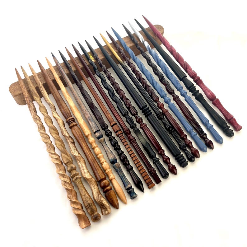 May include: A collection of handcrafted wooden wands in various colors and designs. The wands are displayed on a wooden stand, showcasing a range of styles from twisted to intricately carved. Colors include natural wood tones, black, blue, and red.