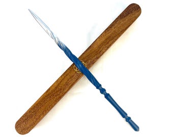 May include: A blue glass wand with a twisted, tapered point that fades to a lighter shade at the tip. The wand is positioned diagonally across a long, rectangular wooden case with a visible grain and a small gold-colored metal ring.