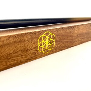 May include: A wooden box with a black and gold pen resting on top. The box is brown with a yellow flower of life symbol on the side. The pen is long and slender.