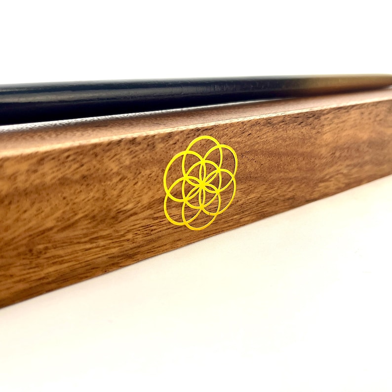 May include: A wooden box with a black and gold pen resting on top. The box is brown with a yellow flower of life symbol on the side. The pen is long and slender.