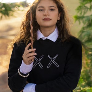 May include: A black sweatshirt with white embroidered "XX" design, layered over a white collared shirt and a white pleated skirt. A wooden wand is held in the hand. A pearl bracelet is worn on the wrist.