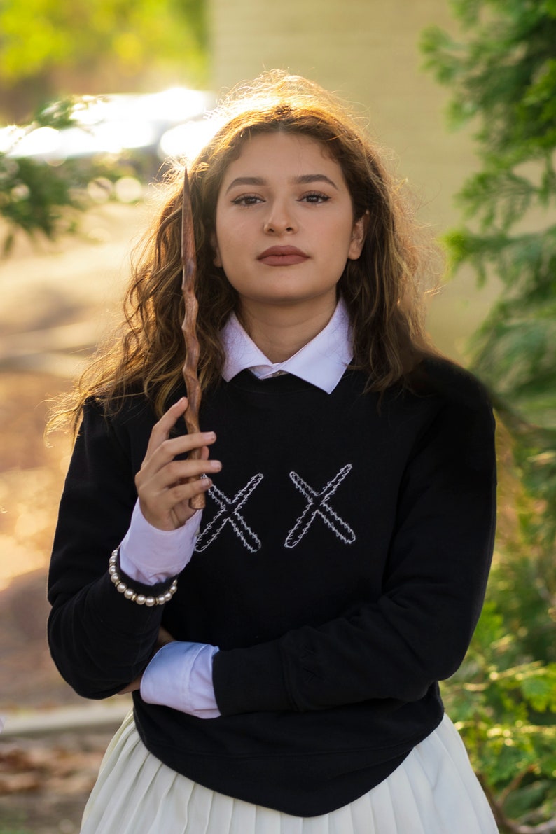 May include: A black sweatshirt with white embroidered "XX" design, layered over a white collared shirt and a white pleated skirt. A wooden wand is held in the hand. A pearl bracelet is worn on the wrist.