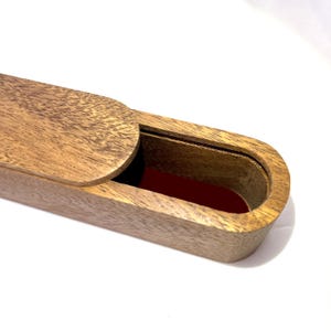 May include: A wooden box with a sliding lid, partially open to reveal a red interior. The box is made of light brown wood with a visible grain pattern. The box is oval-shaped.