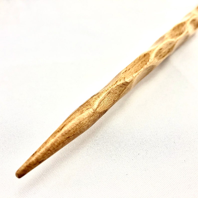 May include: A light brown wooden hair stick with a carved, textured design. The stick tapers to a point at one end, designed for hair styling. The wood grain is visible, enhancing the natural look. A hair accessory.