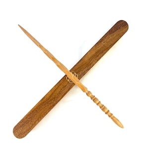 May include: A light brown wooden wand with a carved handle and a pointed tip, resting on a matching wooden case. The wand is angled across the case, with a gold-coloured ring accent. The case is rectangular with rounded ends.