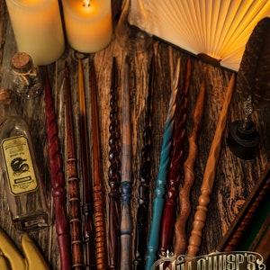 May include: A collection of handcrafted wands in various colors and designs, including red, brown, blue, and black. The wands are arranged on a wooden surface, with candles, a book, and potion bottles in the background. The image is from Willowisp's Wand Emporium.