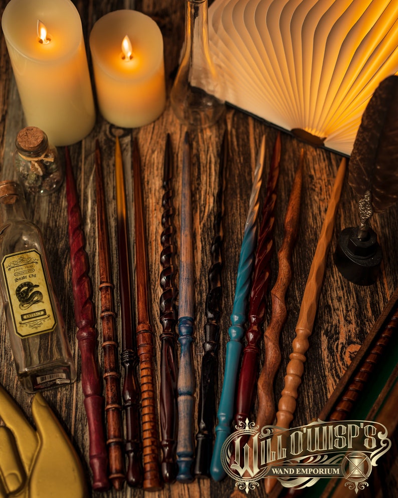 May include: A collection of handcrafted wands in various colors and designs, including red, brown, blue, and black. The wands are arranged on a wooden surface, with candles, a book, and potion bottles in the background. The image is from Willowisp's Wand Emporium.