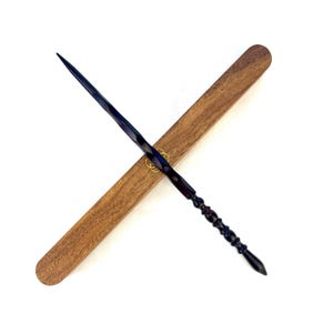 May include: A dark, intricately carved wand with a twisted design, resting diagonally across a wooden case. The wand is dark blue and black, with a detailed handle. The case is a light brown color, with a gold emblem. The wand is approximately 14 inches long.