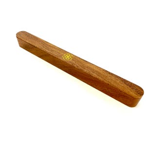 May include: A rectangular wooden box with rounded edges. The box is a warm brown color and has a gold-colored geometric design on the top. The box is long and narrow, and appears to be made of solid wood.