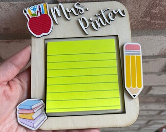 Può includere: Una cornice in legno con un blocco di note adesive giallo, una mela rossa con materiale scolastico, una pila di libri colorati e una matita gialla. La cornice dice "Mrs. Pinter" in alto.