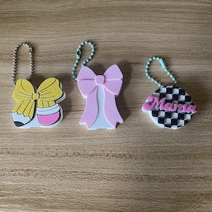 May include: Three decorative keychains with ball chains. The first is a yellow and white pencil-shaped bow. The second is a light pink bow. The third is a circular black and white checkered charm with the pink text Mama written across the center.