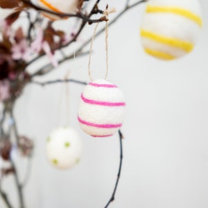 Puede incluir: Un huevo de Pascua de fieltro blanco con rayas rosas colgando de una rama. El huevo est&aacute; decorado con hilo rosa.