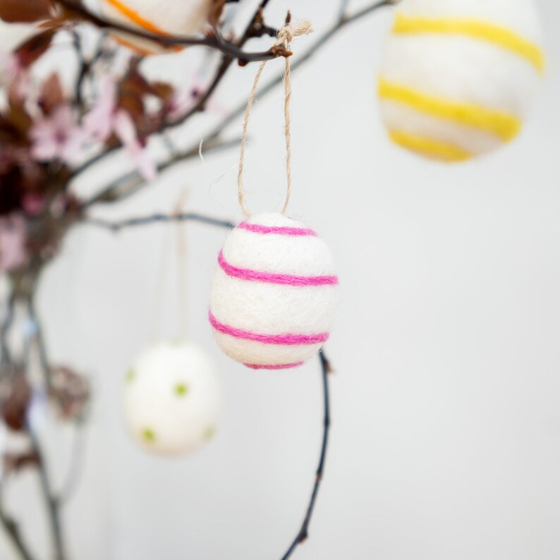 Puede incluir: Un huevo de Pascua de fieltro blanco con rayas rosas colgando de una rama. El huevo est&aacute; decorado con hilo rosa.