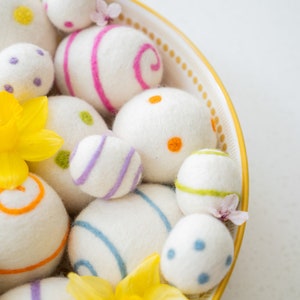 Puede incluir: Un bol lleno de huevos de Pascua de fieltro blanco, algunos con rayas y puntos de colores. Los huevos est&aacute;n dispuestos en un patr&oacute;n decorativo, con algunas flores amarillas asomando por el lado.