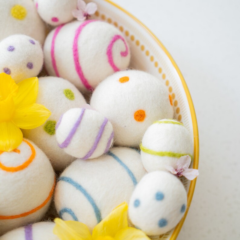 Puede incluir: Un bol lleno de huevos de Pascua de fieltro blanco, algunos con rayas y puntos de colores. Los huevos est&aacute;n dispuestos en un patr&oacute;n decorativo, con algunas flores amarillas asomando por el lado.