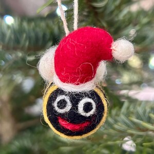 Pode incluir: Um enfeite de feltro preto com um rosto sorridente, olhos brancos e uma boca vermelha. O enfeite está usando um chapéu de Papai Noel vermelho com uma pompom branco.