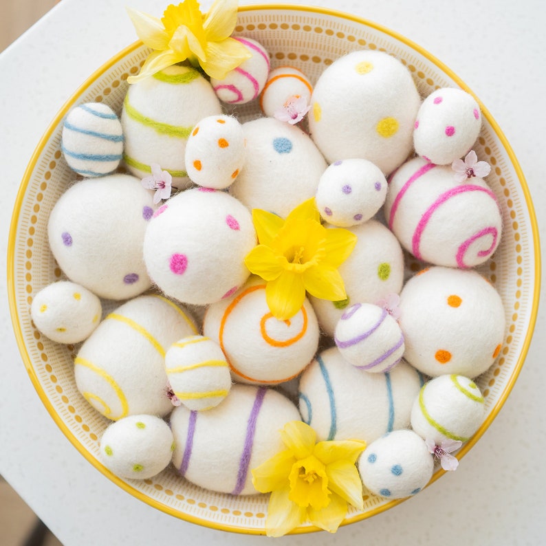 Puede incluir: Un bol lleno de huevos de Pascua de fieltro blanco decorados con rayas, puntos y remolinos de colores. Los huevos est&aacute;n rodeados de narcisos amarillos.