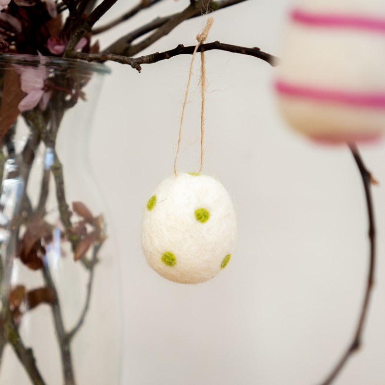 Puede incluir: Un adorno de huevo de Pascua de fieltro blanco con lunares verdes colgando de una rama.
