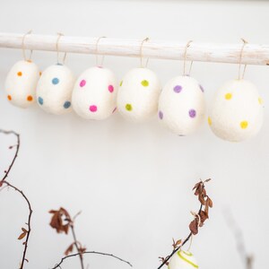Puede incluir: Seis huevos de Pascua de fieltro blanco con lunares de colores que cuelgan de una varilla de madera blanca. Los huevos est&aacute;n decorados con lunares rosas, morados, amarillos, verdes y azules.