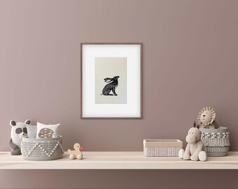 May include: A framed black and white illustration of a hare. The artwork is displayed on a light background within a brown frame. The image is part of a nursery decor setup, with stuffed animals and decorative baskets.