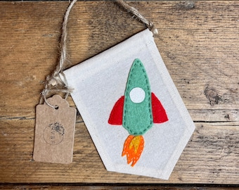 May include: A handmade banner featuring a green rocket ship with red wings and orange flames. The rocket is appliqued on a cream-colored fabric banner, with a jute string for hanging. A small tag with a logo is attached.