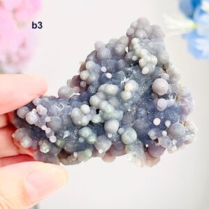 May include: A piece of botryoidal chalcedony in shades of lavender, grey, and white. The surface is covered in small, rounded formations, creating a bubbly texture. The mineral specimen is held in a hand, with the text "b3" visible.