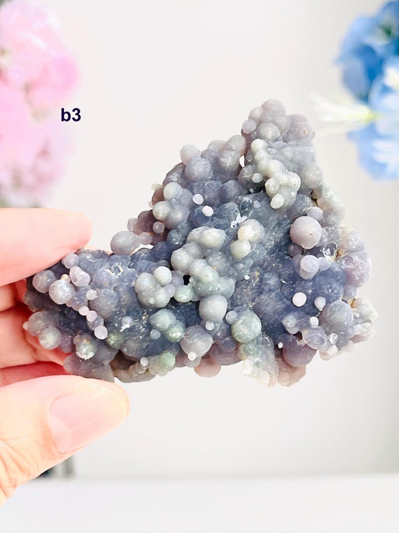 May include: A piece of botryoidal chalcedony in shades of lavender, grey, and white. The surface is covered in small, rounded formations, creating a bubbly texture. The mineral specimen is held in a hand, with the text "b3" visible.