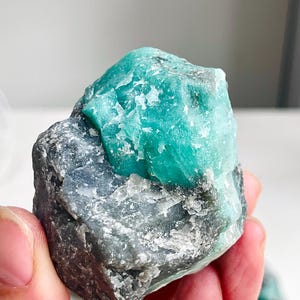 Può includere: Una gemma di smeraldo grezza, non tagliata, di un vibrante colore verde-azzurro, incastonata in una matrice di roccia grigia. Lo smeraldo grezzo mostra una struttura cristallina, con texture e variazioni naturali. La gemma è tenuta in una mano.