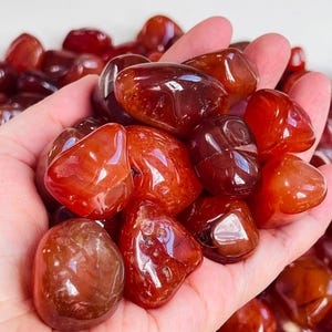 May include: A collection of polished carnelian stones. The stones are a mix of red and orange hues, with a shiny, reflective finish. The gemstones are smooth and rounded, displaying natural colour and texture variations. Suitable for jewellery making or collecting.