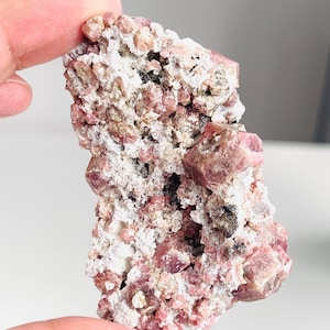 May include: A natural mineral specimen with a mix of pink and white colours. The surface is textured with visible crystal formations and small, embedded pink crystals. The overall appearance is rugged and earthy, with some dark inclusions.
