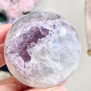 May include: A polished spherical amethyst geode featuring a smooth, rounded exterior and a central cavity filled with deep purple, jagged crystal points. The stone displays a mix of translucent white quartz and vibrant violet amethyst formations.