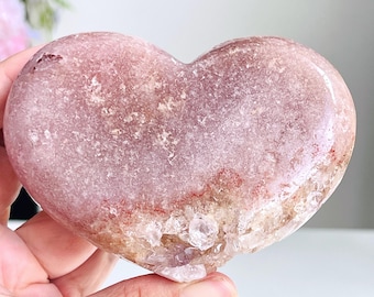 May include: A heart-shaped polished stone featuring a smooth, dusty rose-colored surface with a textured, crystalline base in shades of tan and translucent white. The mineral specimen displays a natural gradient from a solid pink top to a rough, sparkling bottom edge.