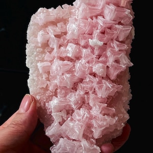 Puede incluir: Primer plano de un grupo de cristales rosas con una superficie rugosa y texturizada. La formación cristalina tiene una apariencia en capas, con numerosos cristales pequeños y angulares. El color varía del rosa pálido a un tono rosa más intenso, con zonas blancas.