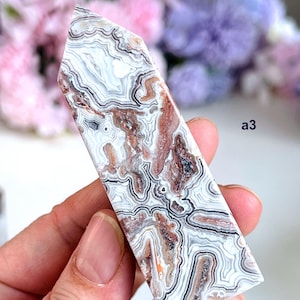 Peut inclure: Une pointe de cristal d'agate polie, présentant un motif de couches blanches, grises, marron et noires. Le cristal est tenu dans une main, avec un fond floral flou. Le texte "a3" est visible sur le côté droit de l'image.