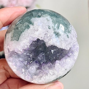 May include: A polished spherical mineral specimen featuring a deep purple amethyst geode centre surrounded by white crystalline structures and patches of dark green moss agate. The surface is smooth and rounded, showcasing natural mineral formations.