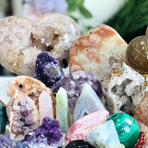 Pode incluir: Uma coleção de vários cristais e pedras preciosas, incluindo uma grande ametista em forma de coração, uma ágata rosa, uma esfera de malaquita verde e vários aglomerados de ametista menores. Os cristais estão dispostos sobre uma superfície branca.