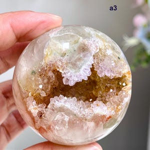 Könnte beinhalten: Ein polierter kugelförmiger Kristall mit einer zentralen Geodenhöhle, die hellviolette Amethystspitzen und goldbraune Mineralablagerungen aufweist. Die Außenfläche ist glatt und durchscheinend, mit dem Text a3 in der oberen rechten Ecke.