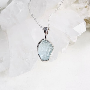 Puede incluir: Un collar de plata con un colgante de cristal de aguamarina azul claro. El cristal es en bruto y tiene una forma natural irregular.