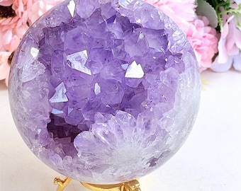May include: A spherical amethyst geode featuring deep purple crystalline structures within a hollow center, supported by a polished gold-toned metal tripod stand. The crystal sphere displays natural variations in color and texture.