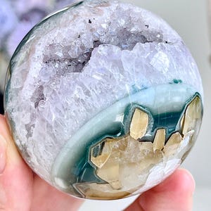 May include: A polished spherical mineral specimen featuring a central geode cavity filled with light purple amethyst crystals, surrounded by layers of dark teal and translucent white agate with embedded golden-yellow crystalline inclusions.