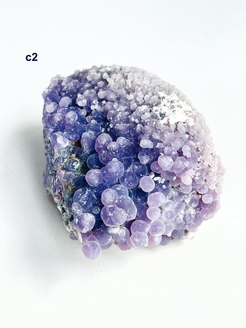 May include: A grape agate specimen, a mineral with a distinctive appearance. It features numerous small, spherical formations, resembling grapes, in varying shades of purple and lavender. The text "c2" is visible.