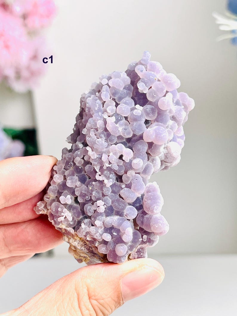May include: A cluster of grape agate, a gemstone with a bubbly, botryoidal structure. The stone has a soft, lavender hue with rounded formations. The base of the stone is a mix of brown and tan. The image also shows the text "c1".